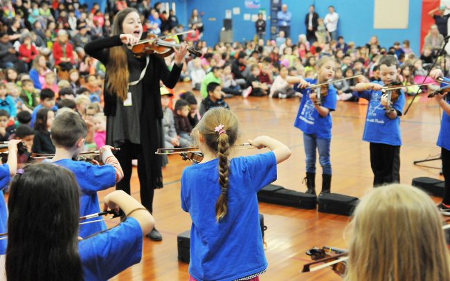 Children playing violins at a school assembly with their instructor