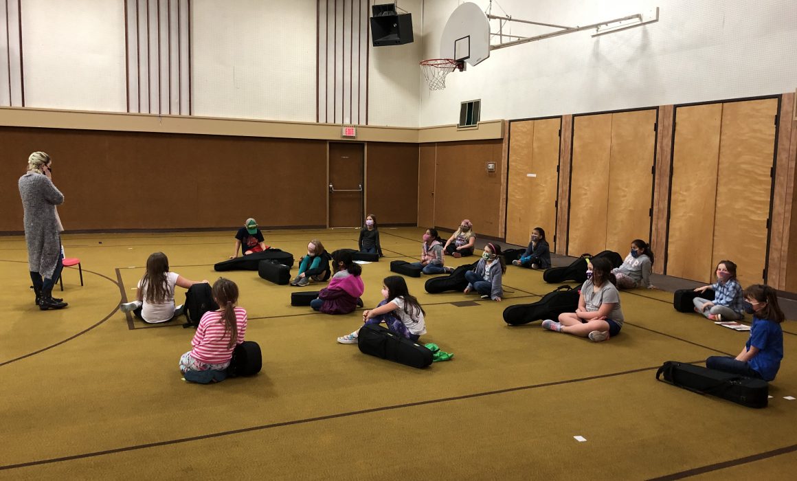 Children seated on the floor of a gymnasium next to violins